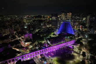 Vista aérea noturna dos Arcos da Lapa iluminados em tons de rosa e roxo, com a Catedral Metropolitana ao fundo.