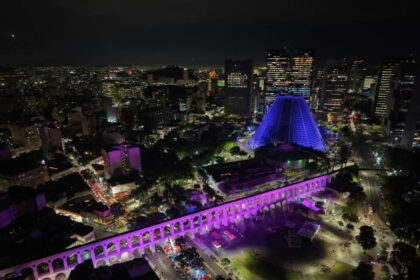 Vista aérea noturna dos Arcos da Lapa iluminados em tons de rosa e roxo, com a Catedral Metropolitana ao fundo.