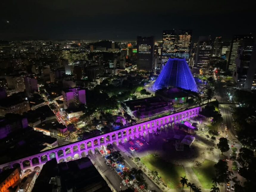 Vista aérea noturna dos Arcos da Lapa iluminados em tons de rosa e roxo, com a Catedral Metropolitana ao fundo.