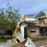 Construção de alvenaria com parte da cobertura arrancada e telhas metálicas espalhadas em frente ao imóvel.